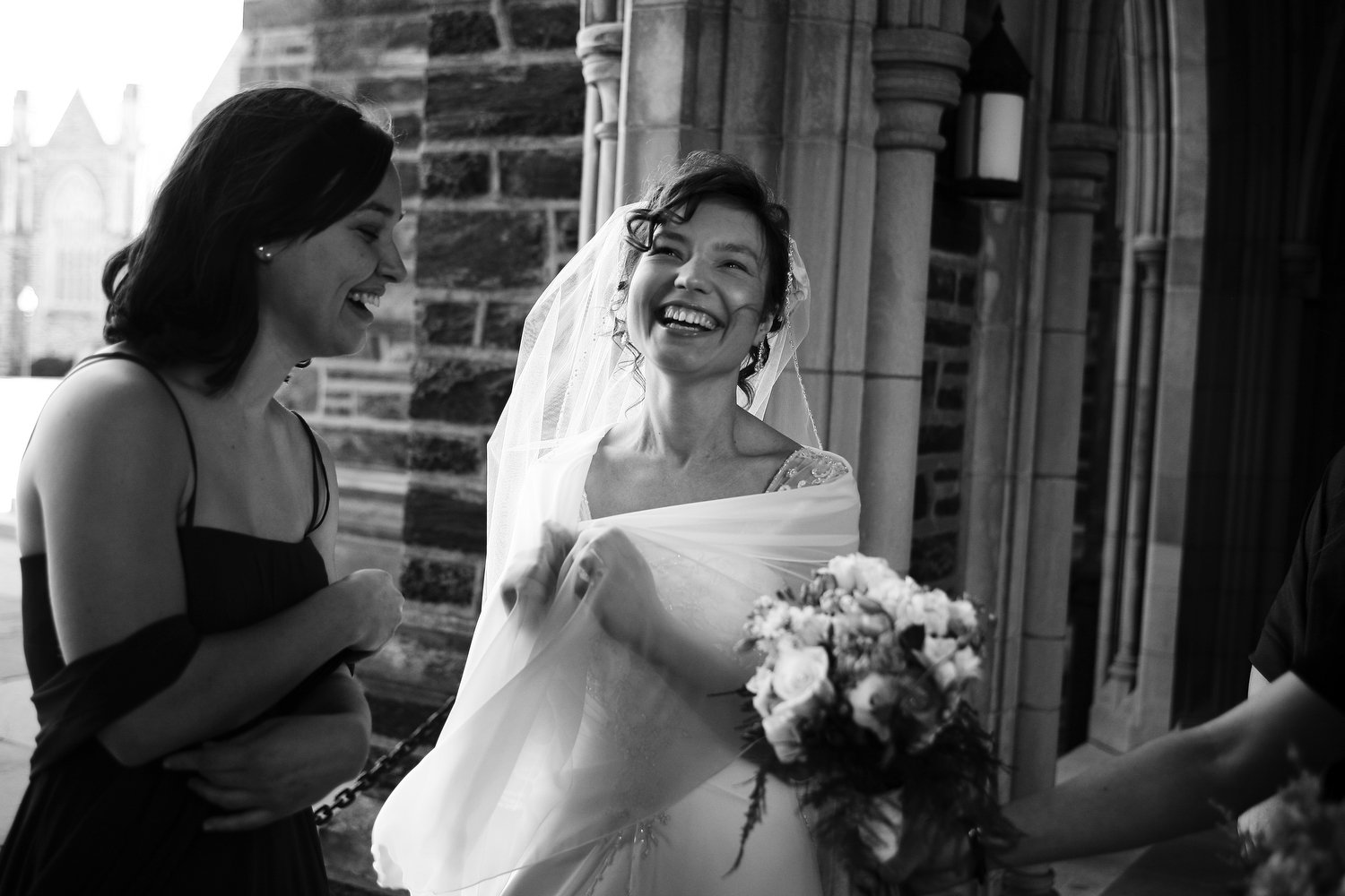 Black and white photo of a bride in a wedding dress clutching her long veil with both hands in front of her and smiling. She is outside a church with stone architecture and a bridesmaid holds a bouquet out to her as another bridesmaid looks on and sm