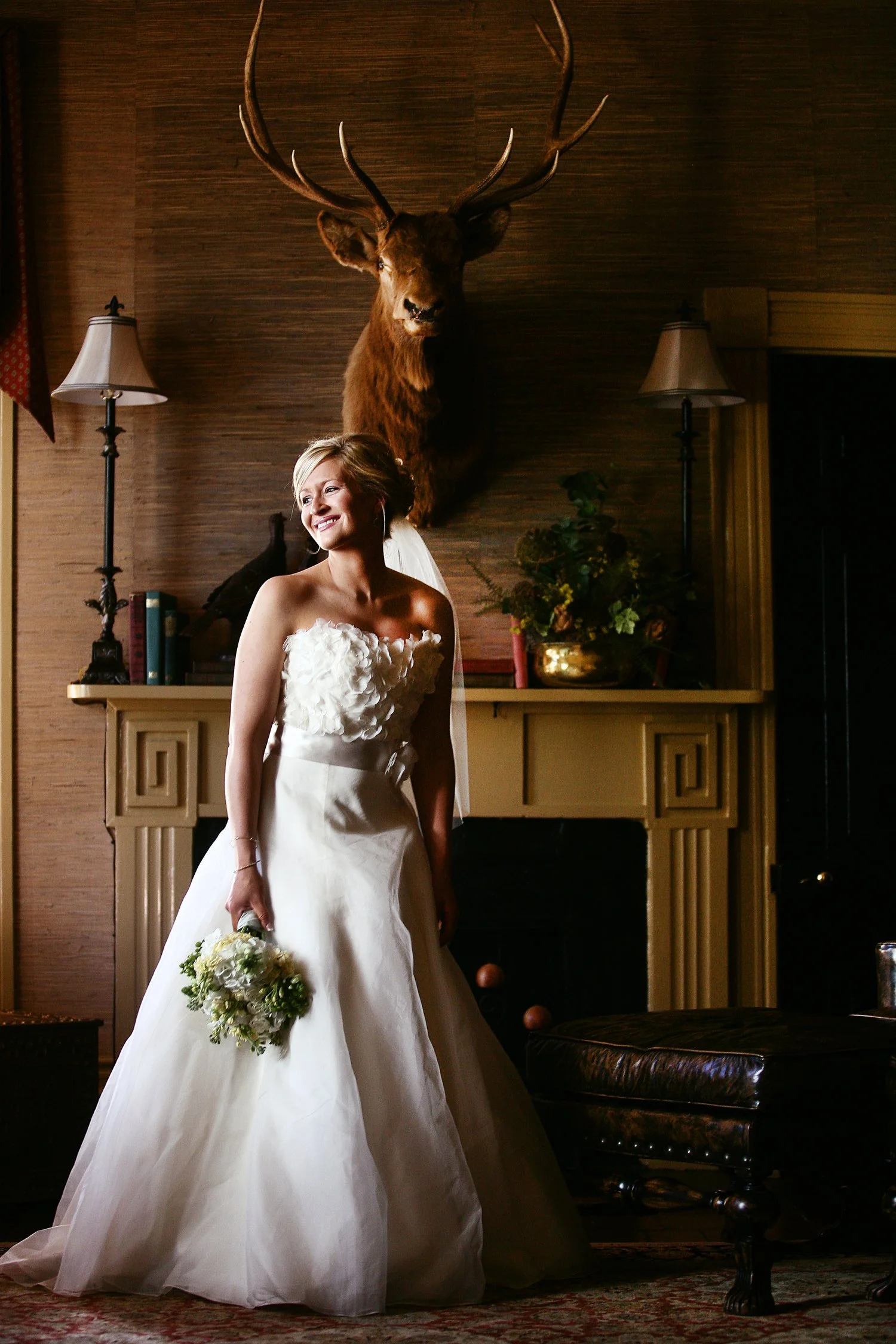 A bride in a strapless white wedding gown clutches a bouquet of flowers in her right hand hanging at her side. On the wall behind her is a large deer head framed with a lamp on either side.
