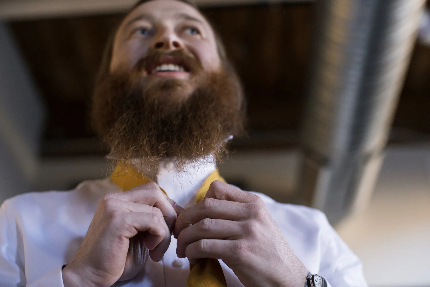 A man with a red beard and mustache adjusting his yellow tie, looking upward with a smile, wearing a white shirt with a yellow collar, in an indoor setting with an industrial pipe in the background above him. Photo is taken from a low angle.