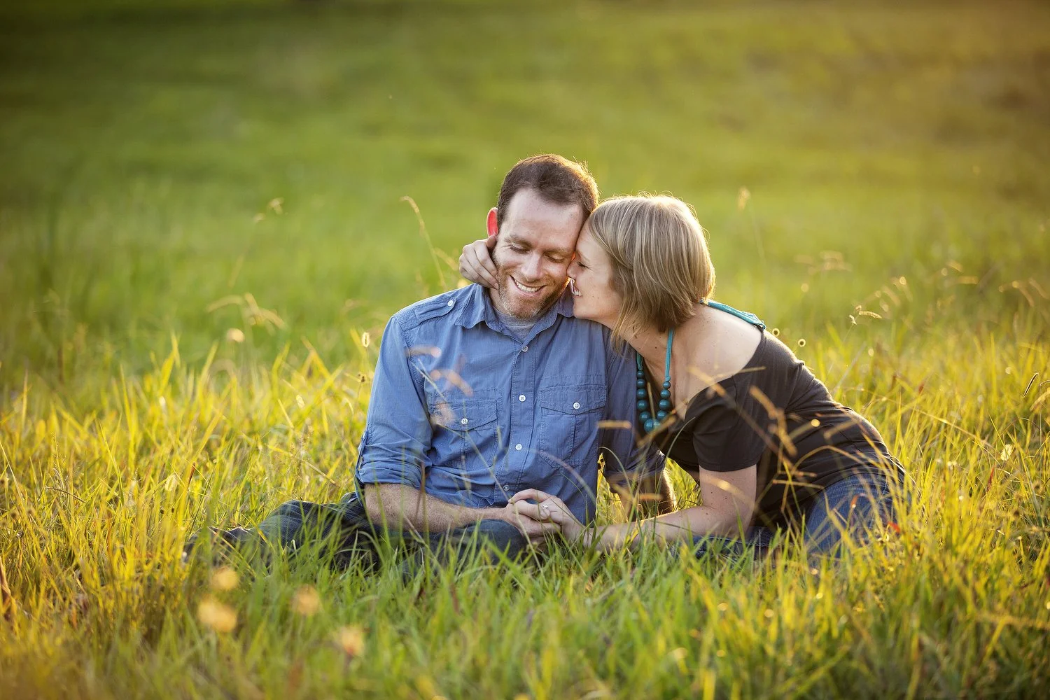 A couple sits together in a wild grassy green field at sunset. The warm glow of the sun surrounds them. She leans in from the side and holds his face in one hand and his hand in the other. She has on a brown shirt and a teal beaded necklace. Her hair