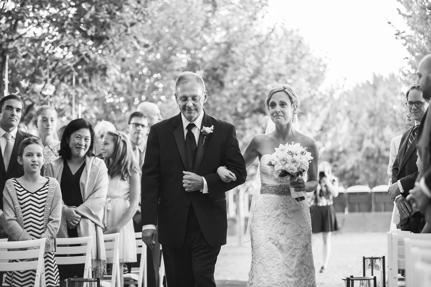 A bride walking down the aisle arm-in-arm with her father during her wedding ceremony. She holds a bouquet of flowers and looks emotional. Guests are visible in the background, watching the procession. The photo is black and white. 