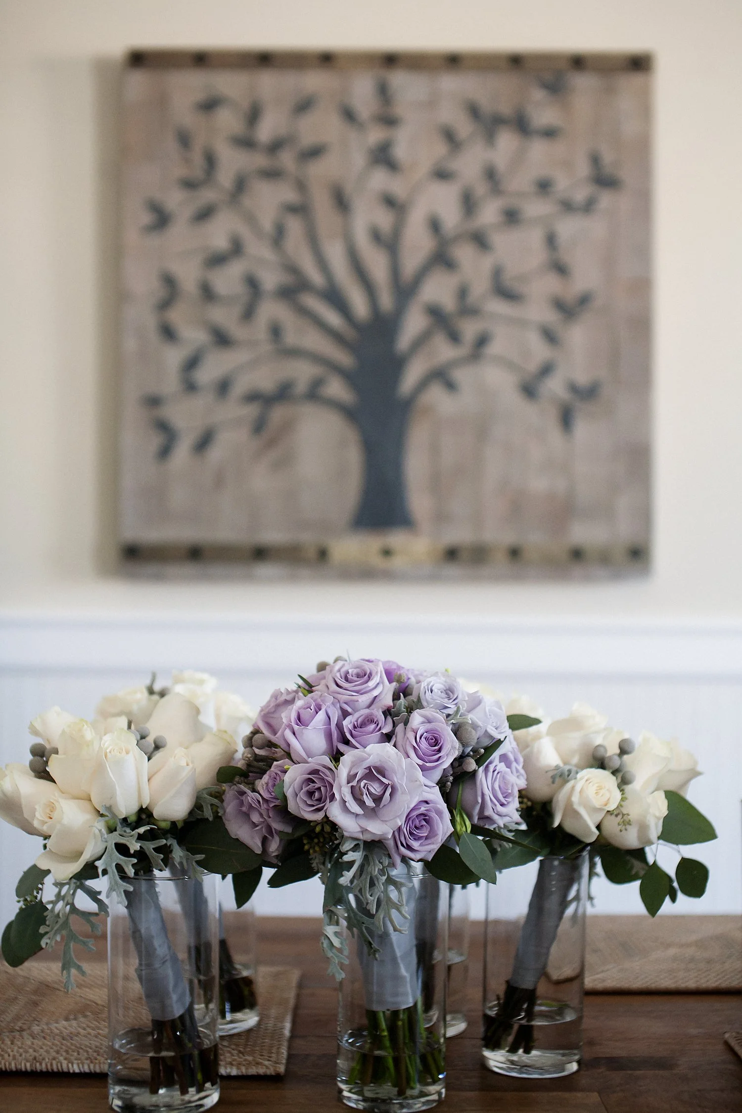 Three bouquets of roses sit in glass jars of water on a wooden table. There is a decorative piece of art with a tree on it on the wall behind the flowers. The middle bouquet is a lavender color. The ones on either side of it are white. 
