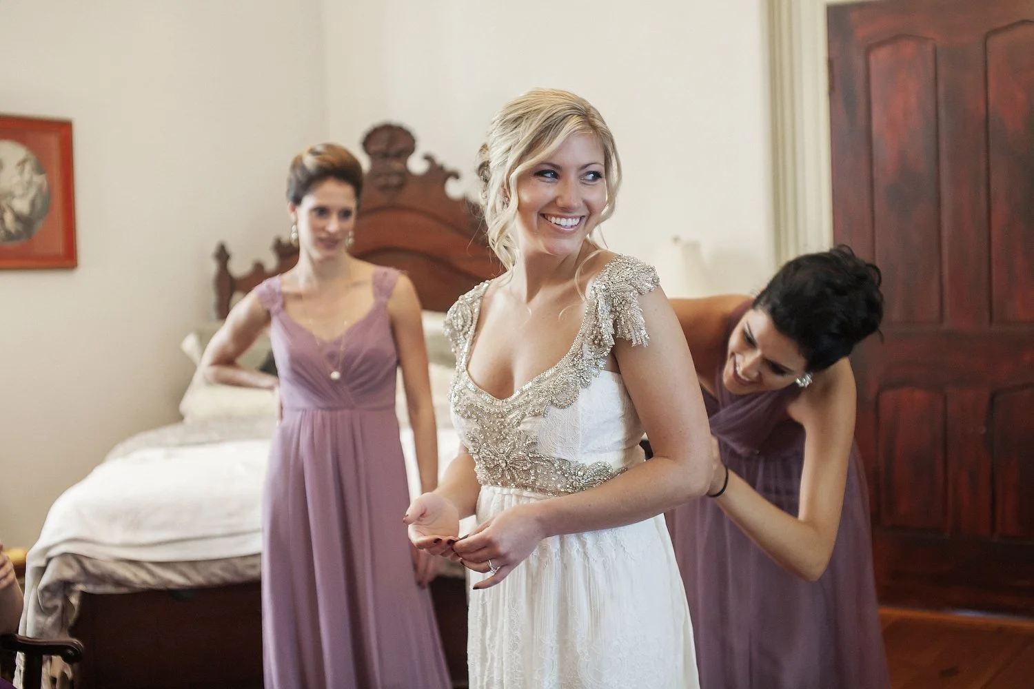 A bride in a white vintage wedding dress with beadwork is smiling as she stands in a bedroom, with two bridesmaids dressed in mauve dresses helping her get ready.