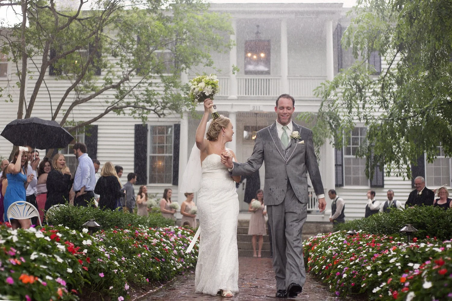 A newlywed couple walks arm-in-arm in the rain outdoors, with the bride holding a bouquet up in the air. Both are smiling, surrounded by friends and family under umbrellas, in front of a large white house with flower beds along a brick path.
