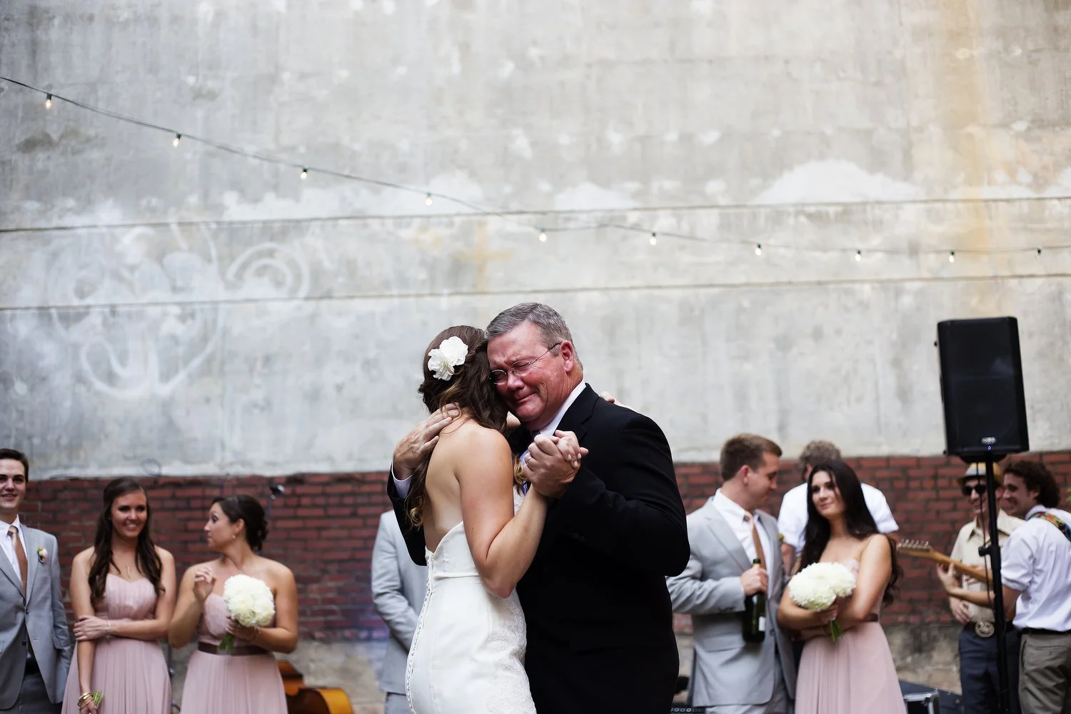 Outdoor reception venue next to a building. There is a cement wall with a brick bottom. String lights are seen above, but it's still light outside. The wedding party, men in grey suits and women in pink dresses, stand in the background as a bride dan