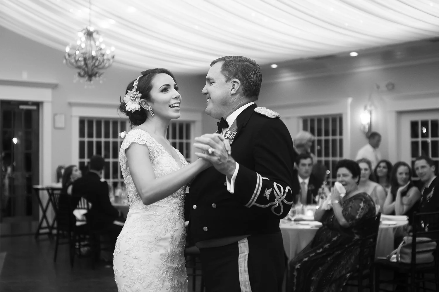 A bride in a lace wedding dress dancing with a man in a military uniform at a reception with guests seated at tables in the background.