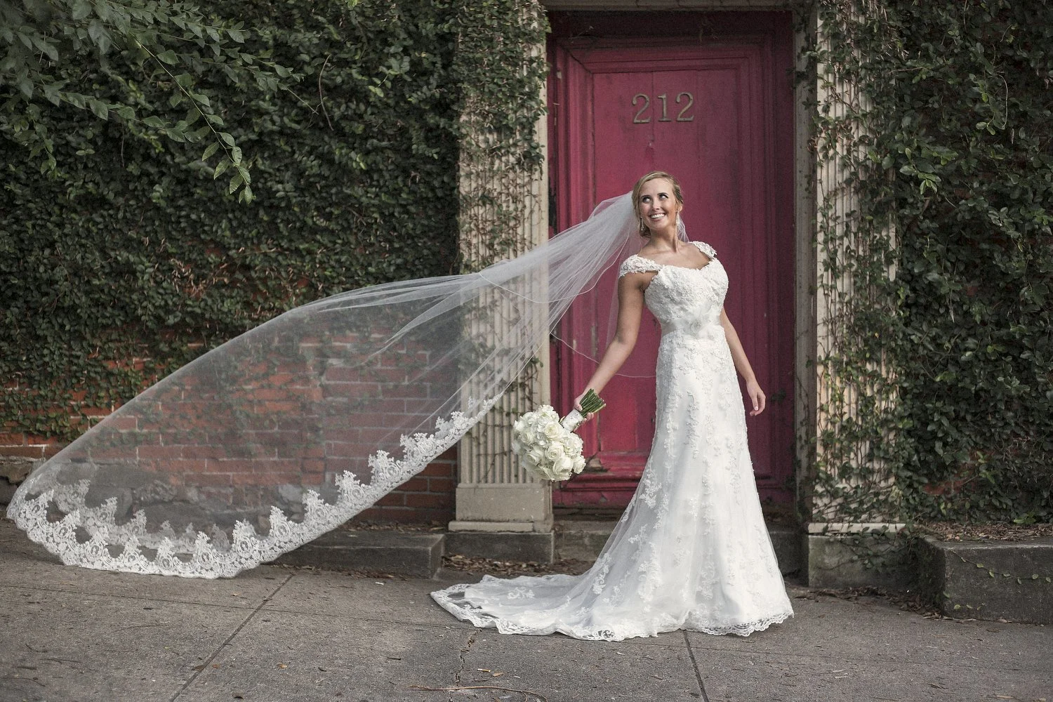 A bride in a white wedding dress holding a bouquet of white roses standing outside near a bright pinkish-red door with the number 212, surrounded by green ivy. Her floor length veil is blowing out beside her.