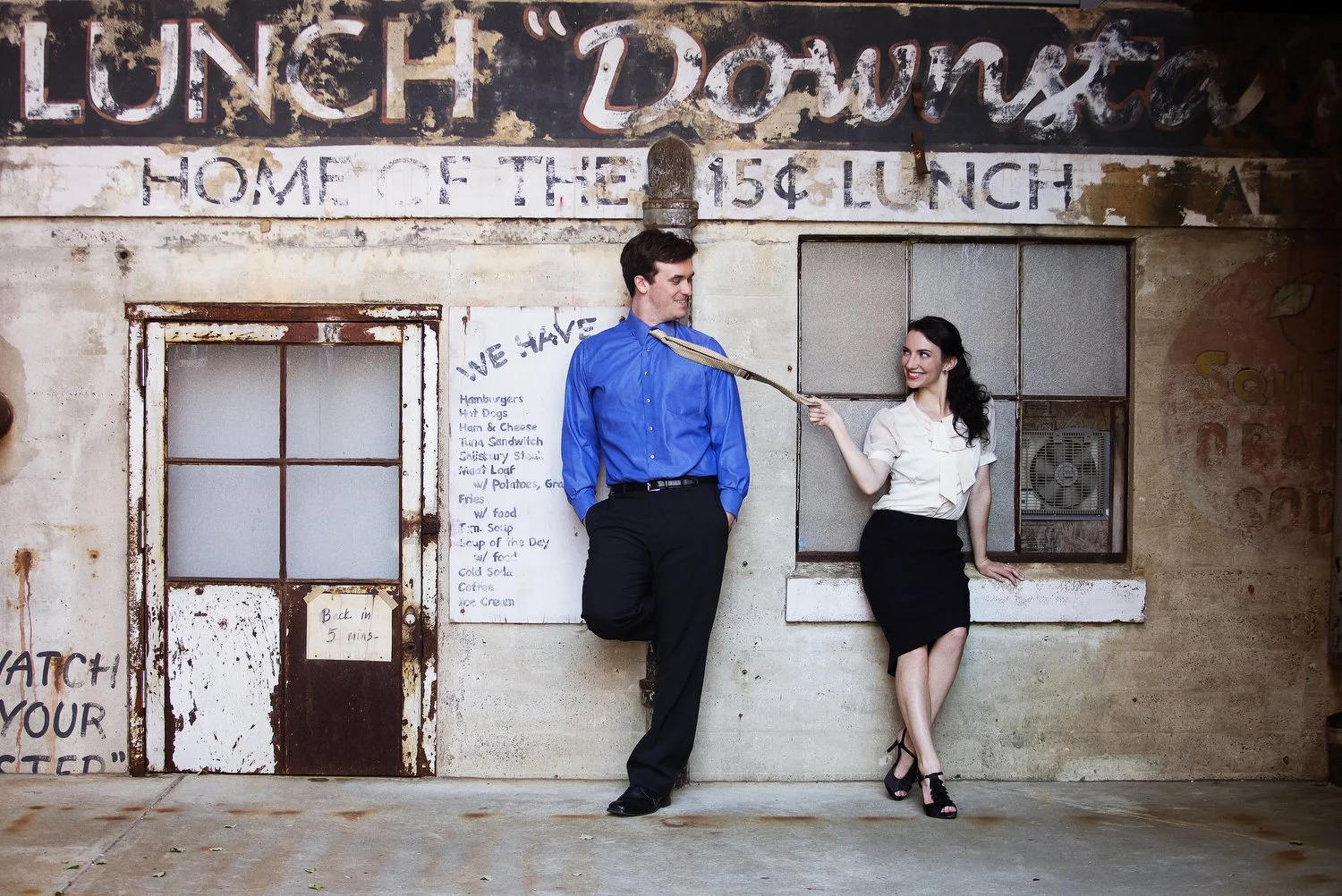 Creative couples portrait in front of a rustic vintage storefront at the Tobacco Campus in Durham, North Carolina with faded signage reading 'Lunch Down Home Since 1954." Photographed in warm natural light, this image captures the playful chemistry a