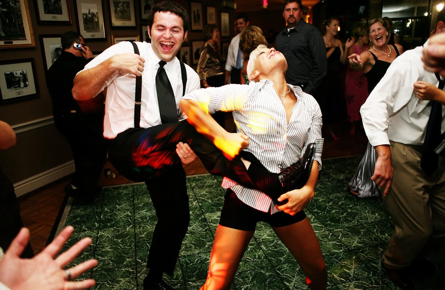 A young man and woman are seen at the center of a green dance floor. Pictures hang on the wall behind them. Other people are dancing off to the sides and looking on from behind them. The woman is wearing short black shorts and a white button up shirt