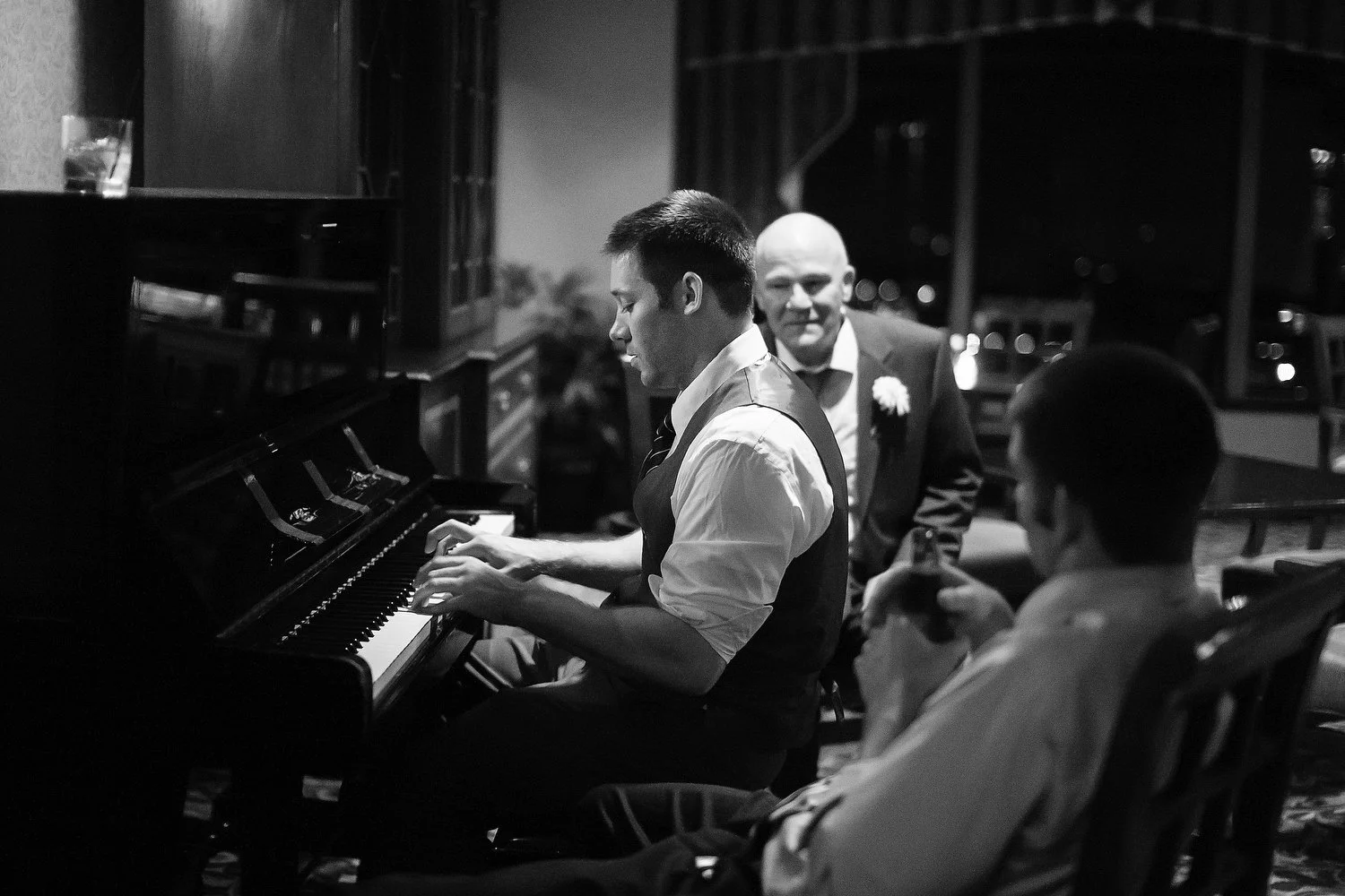 A black and white photo of a young man playing piano at an event, with an older man in a suit and boutonniere and a woman in glasses and a dress watching him.