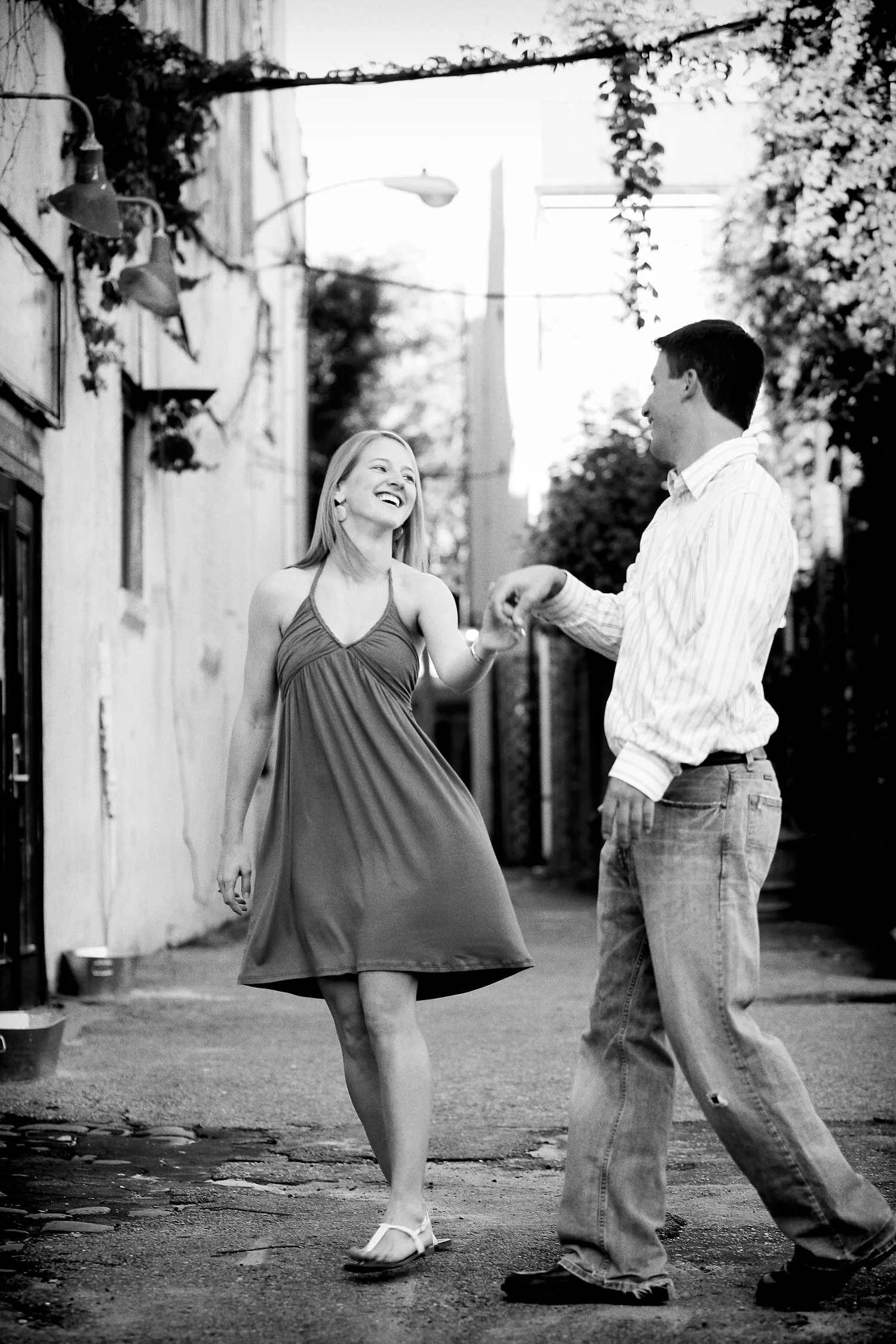 A couple stands together in a cobblestone street in between old brick and stone buildings. Some vines stretch from one building to the other up above them. She wearing a cotton dress with spaghetti straps that is knee-length. He has on a white dress 