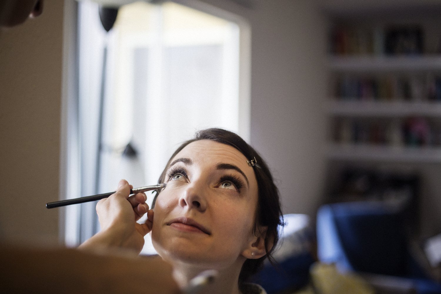 Woman with dark hair and blue eyes having her makeup applied, looking up while a makeup artist uses a brush near her eye.