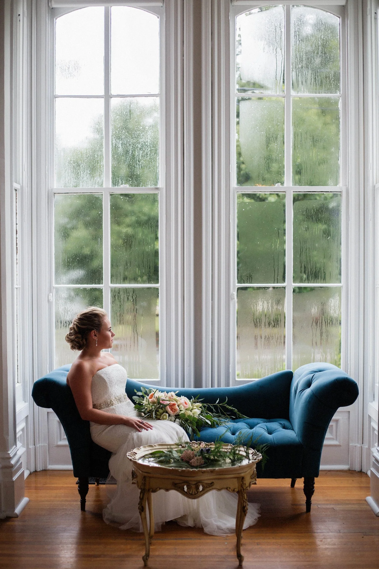 A bride sitting on a blue velvet chaise lounge in front of large rain-beaded windows, wearing a strapless white wedding dress, with a floral bouquet on her lap and an ornate gold side table in front.