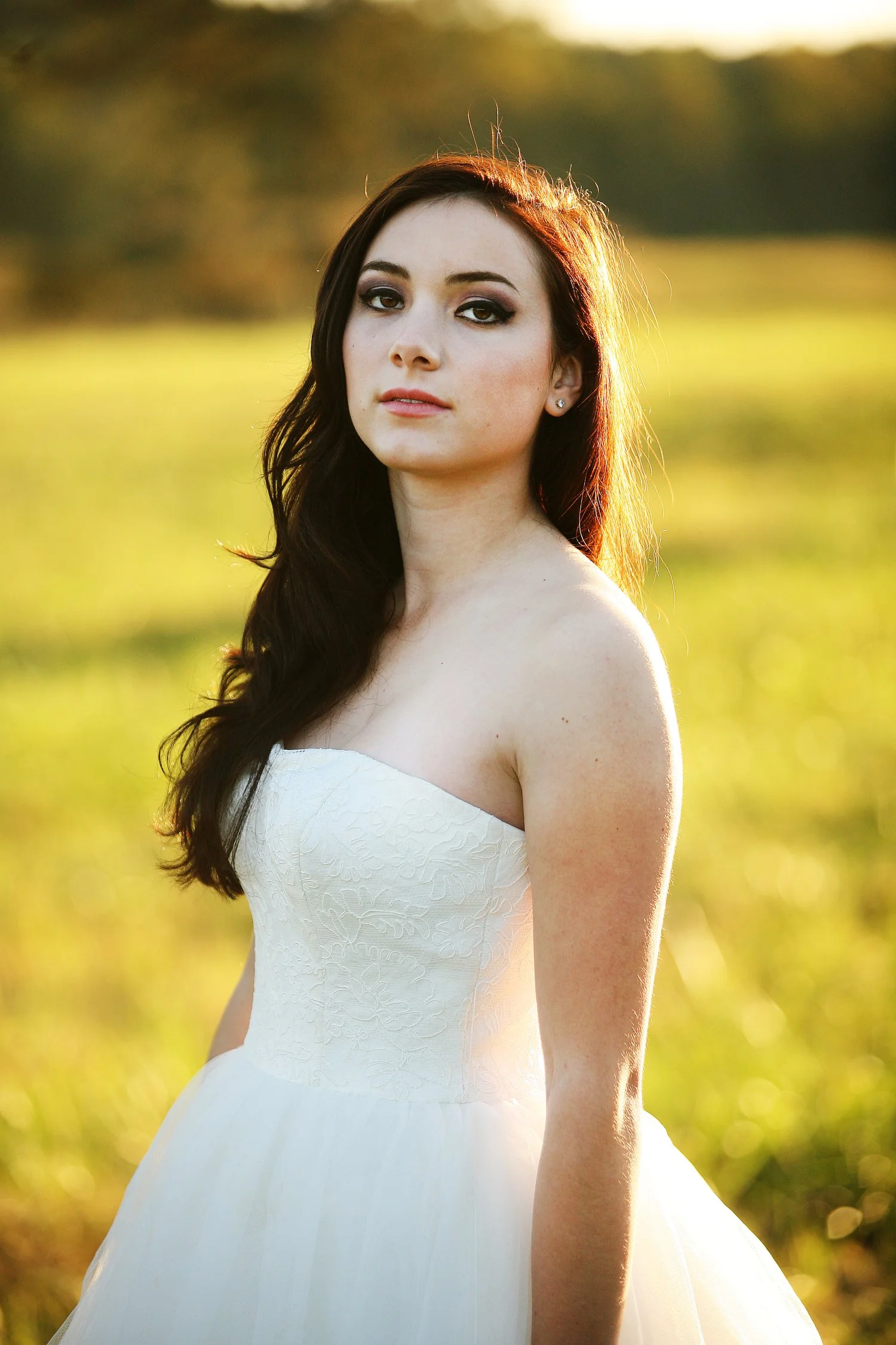 A young woman in a white wedding dress standing outdoors in a golden grassy field during sunset. Her reddish brown hair is down and the sunset light gives it a glow from behind. She has dark makeup on her dark brown eyes and a reddish brown lipstick 