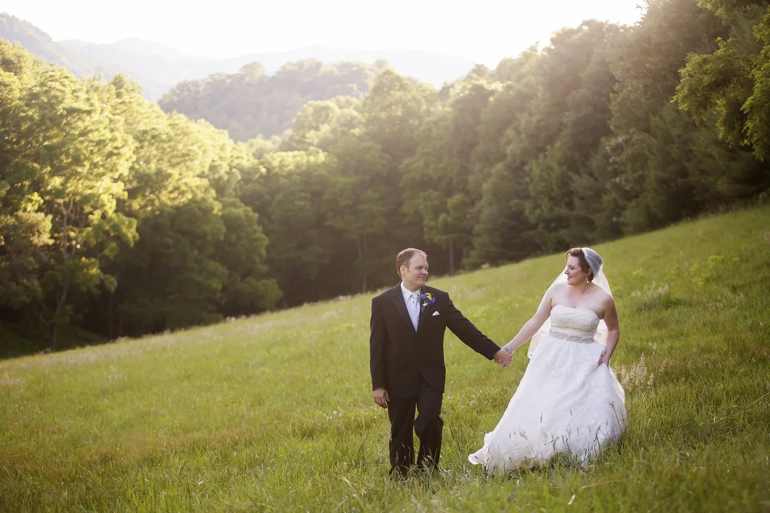 A bride and groom holding hands in a grassy field surrounded by trees and mountains during sunset.