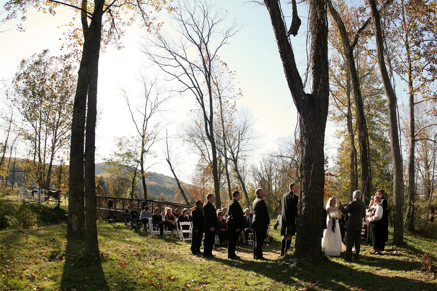 A wedding ceremony outdoors in a wooded area during autumn, with guests seated and the bride and groom standing with an officiant. The view is taken from the distance and is a scene setter of the ceremony.