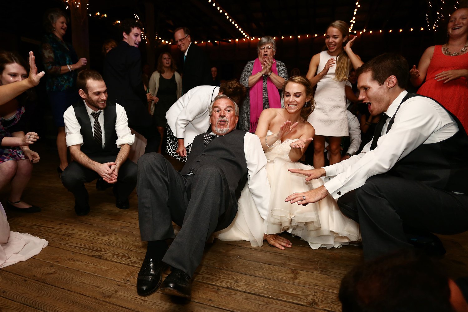 Reception dance floor with the bride in a strapless white dress is down low in the center of the frame. She is smiling and her hands are coming together like she's clapping. An older man is down low next to her in a black suit with a vest and white b