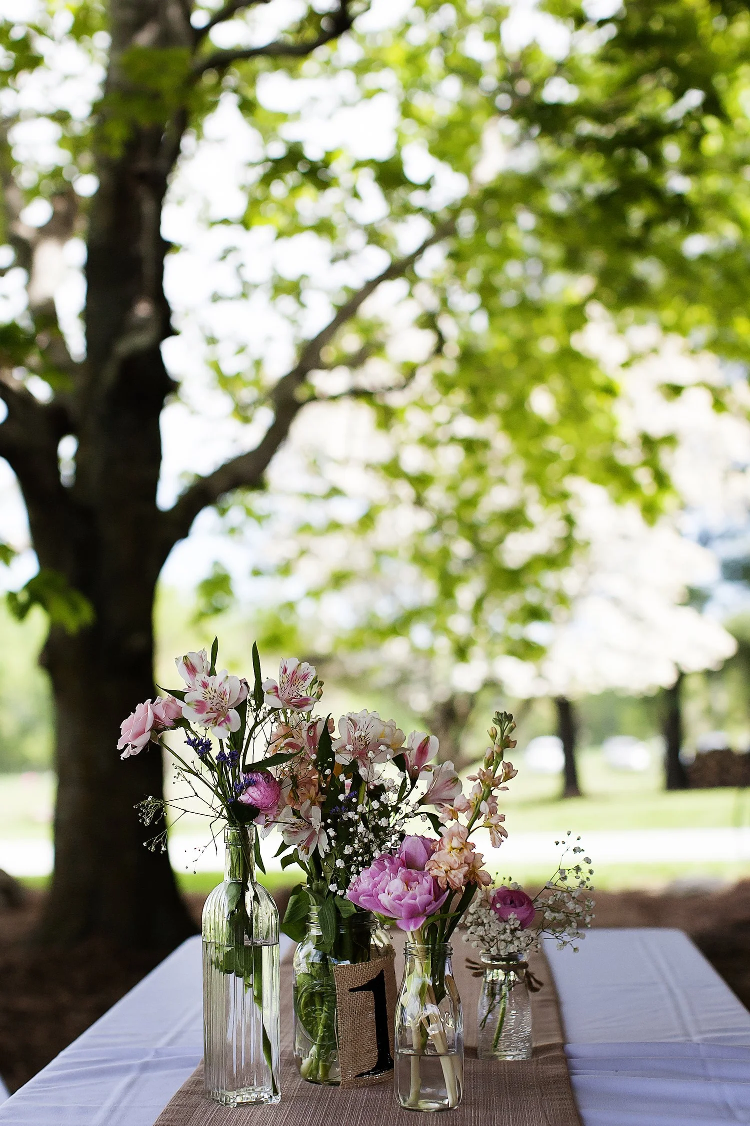 Pink, purple and white flowers are arranged in small glass jars of various sizes on a burlap table runner on a rectangle table with a white table cloth. They are outside and a tree with green leaves frames them in the distance.