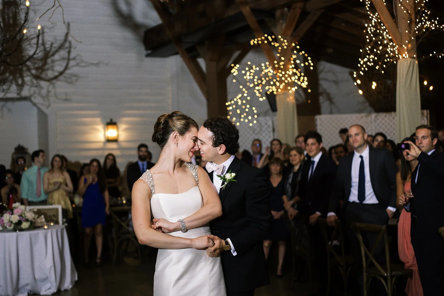 A bride leans in to her groom and their foreheads touch in a tender moment before he appears to be ready to spin her as they dance with their wedding guests behind them. Her white dress has jeweled thick straps on her shoulders. Her hair is up. He ha