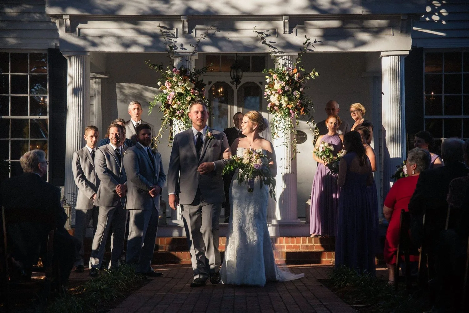 A wedding ceremony taking place outdoors on a porch with a floral arch, with the bride and groom walking down the aisle, surrounded by bridesmaids and groomsmen, with seated guests watching. This is the Leslie Alford Mims House in Holly Springs, Nort