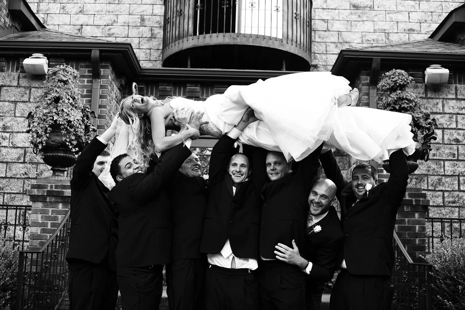 Black and white photo of a group of men in tuxedos lifting a woman in a wedding dress in the air outside of a brick building with a rounded balcony and potted plants.