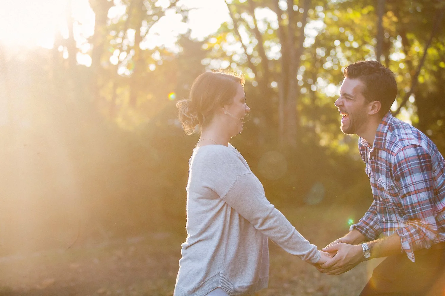 A couple stands in a wooded area at sunset as the warm golden light surrounds them. She has on a long-sleeve shirt and her hair is pulled up in a messy bun. She holds his hands and faces him. He has on a checkered print button-up shirt and is leaning