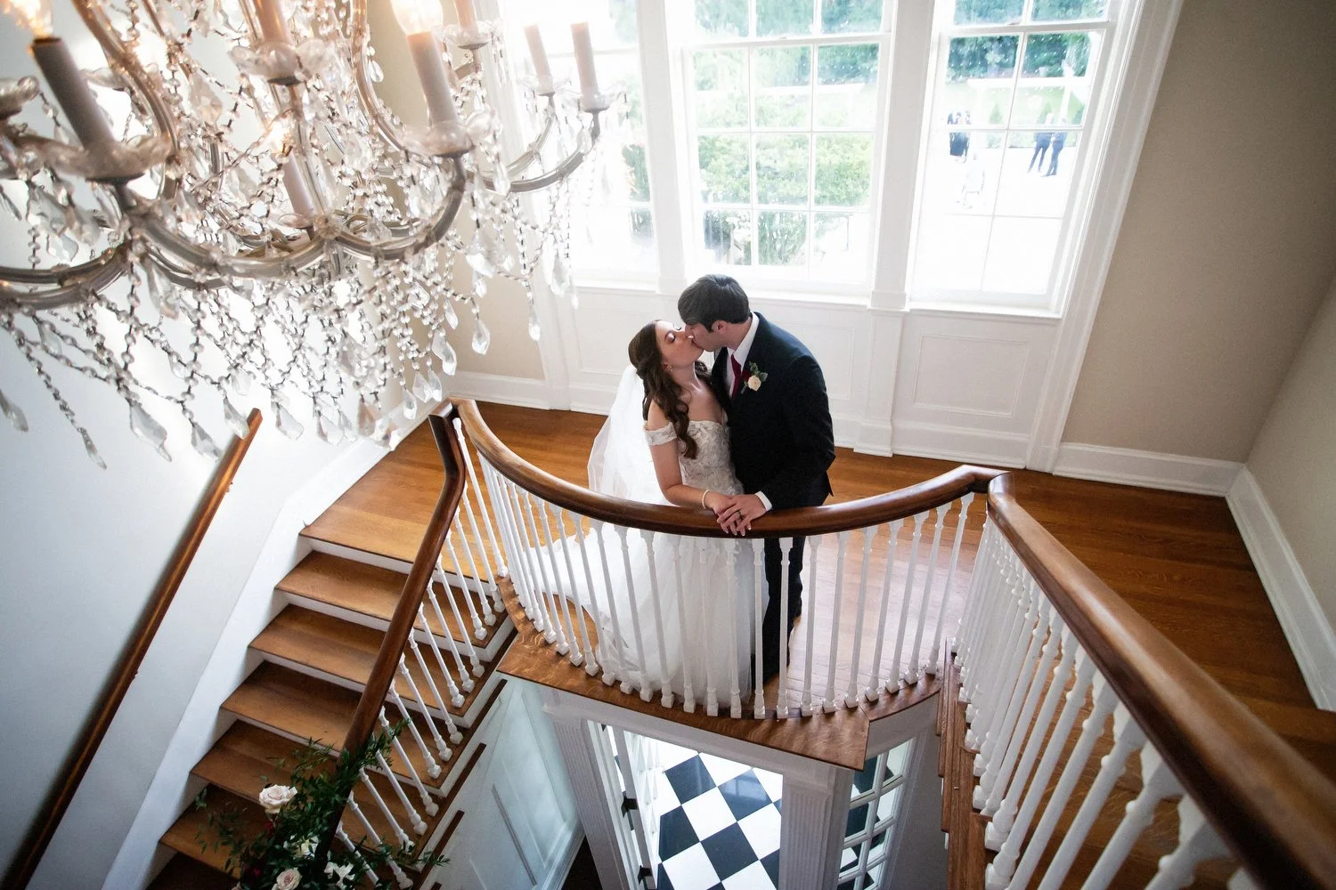 Photo of a bride and groom kissing at a balcony on the landing of a staircase. The photo is taken from the top. A fancy chandelier hangs up above them and near floor length windows with white trim are behind them. He is wearing a black tuxedo and she
