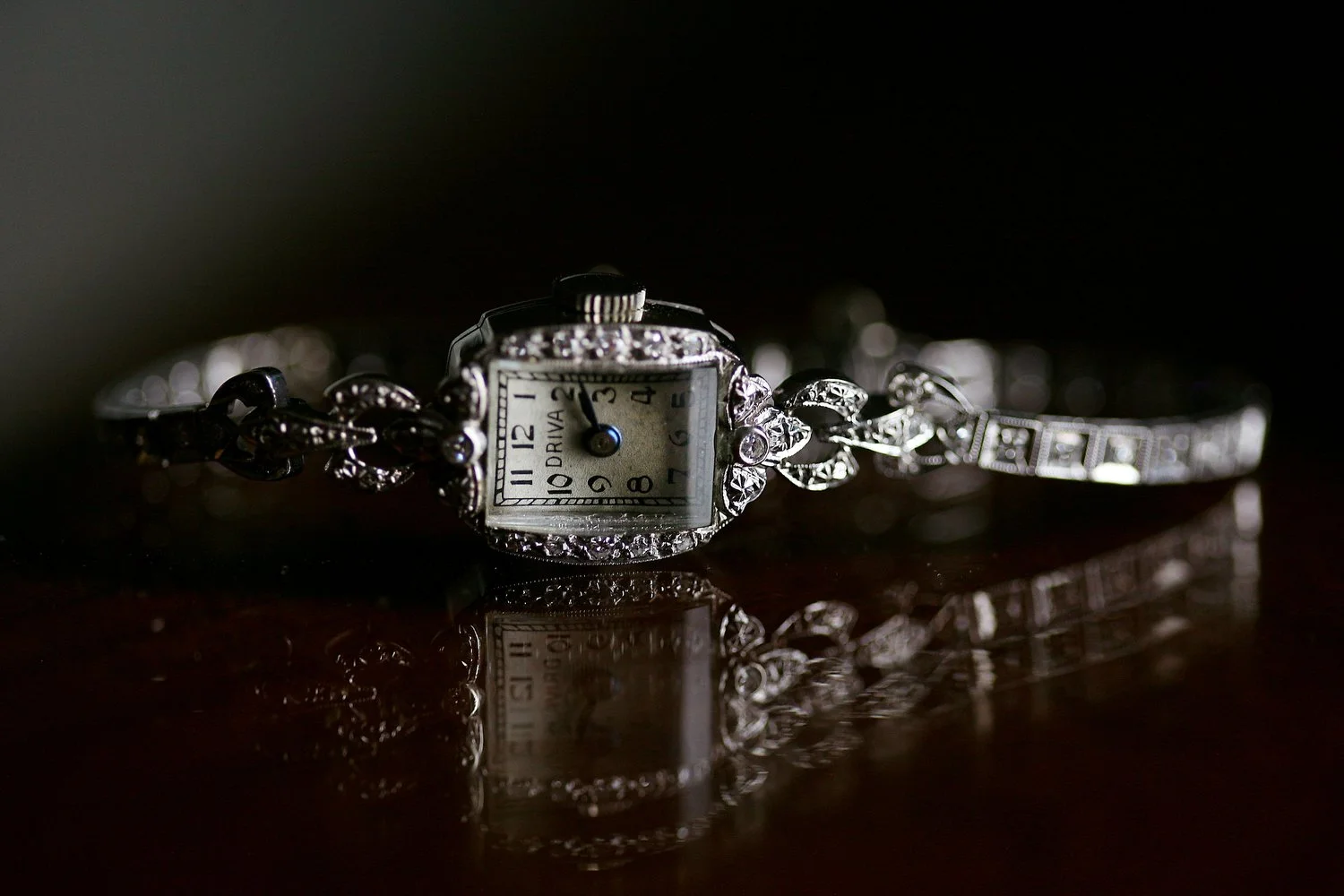 Close up image of a vintage silver watch on a dark brown surface. The window light allows for a reflection of the watch on the table surface. 