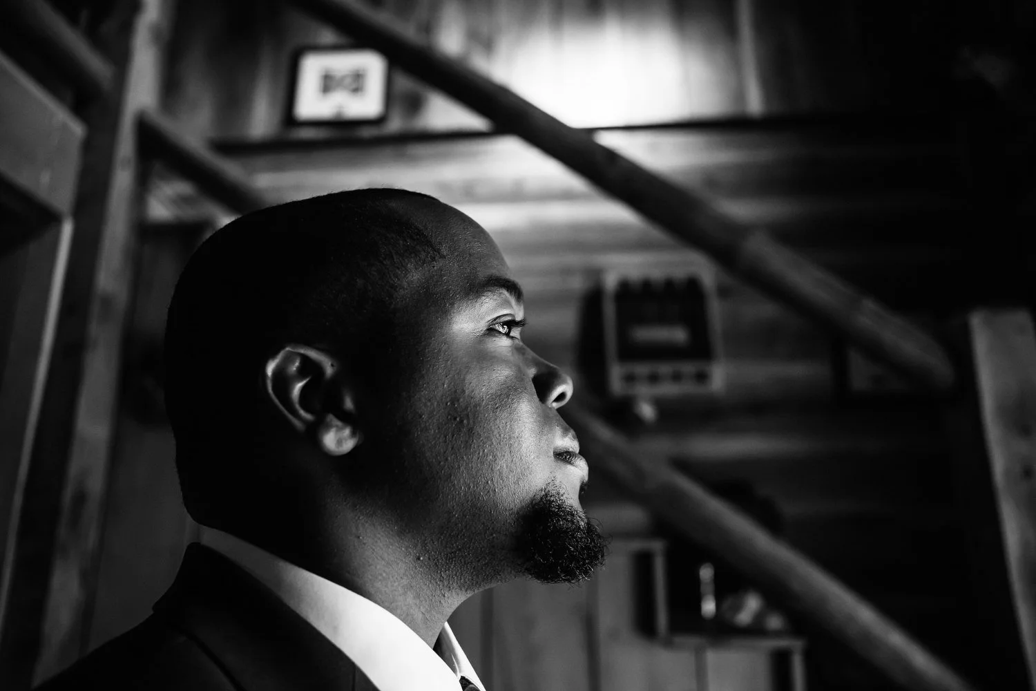 A black and white photo of a man in profile, wearing a suit, looking contemplatively upward indoors with a wooden staircase in the background.