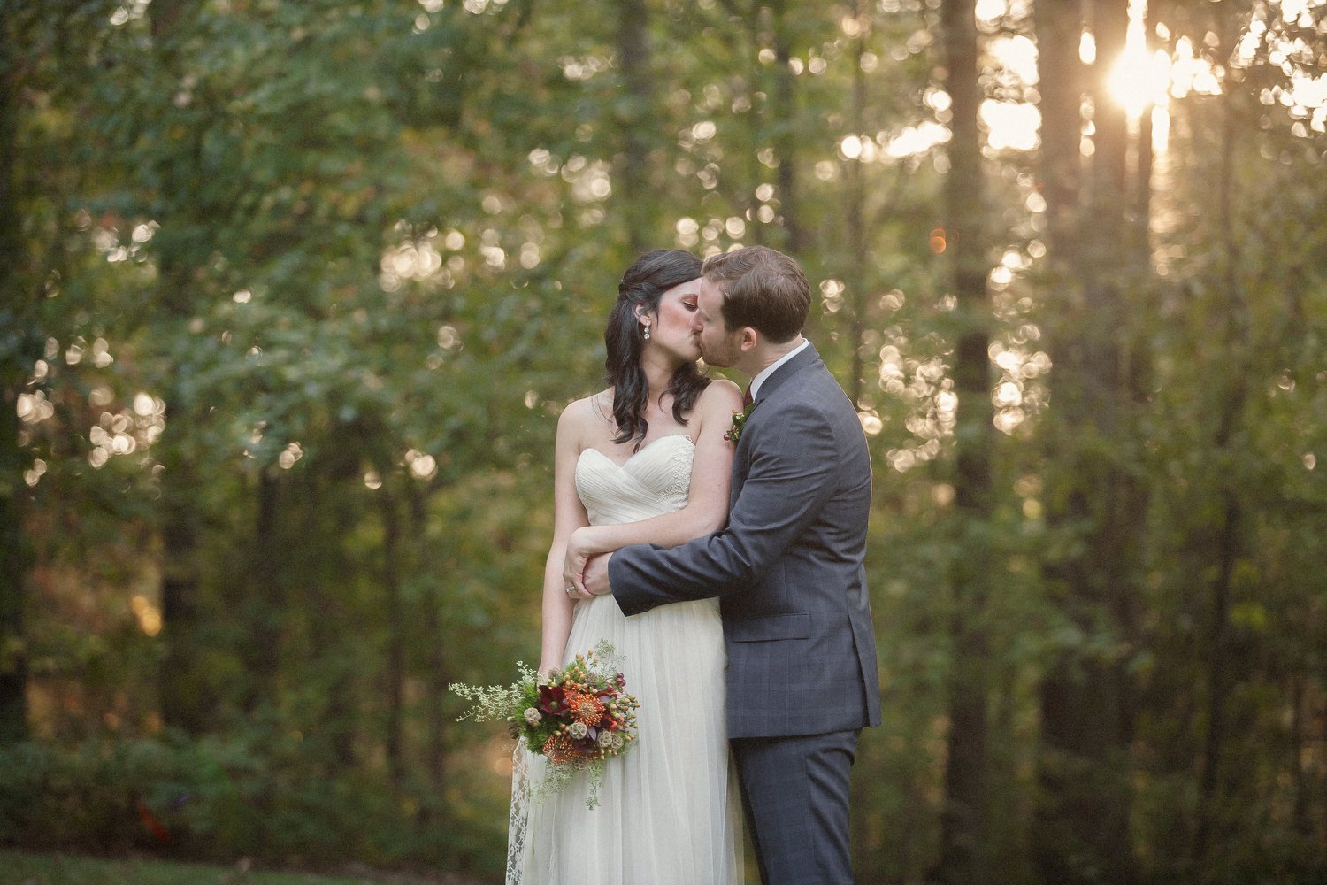 A bride and groom kissing in a wooded outdoor setting during sunset, with the bride holding a bouquet of flowers. The groom has his arm around her waist.