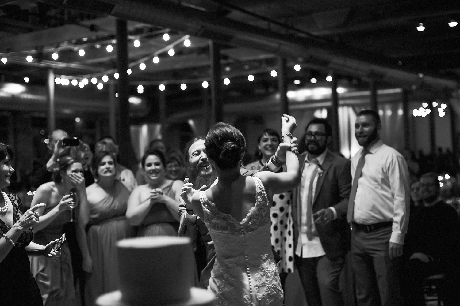 Fun black and white image of the bride and groom playfully feeding each other cake. The cake is in the foreground as the bride is seen from the back with her arm up high trying to put cake on her groom's face. He is grinning and trying to keep her ha