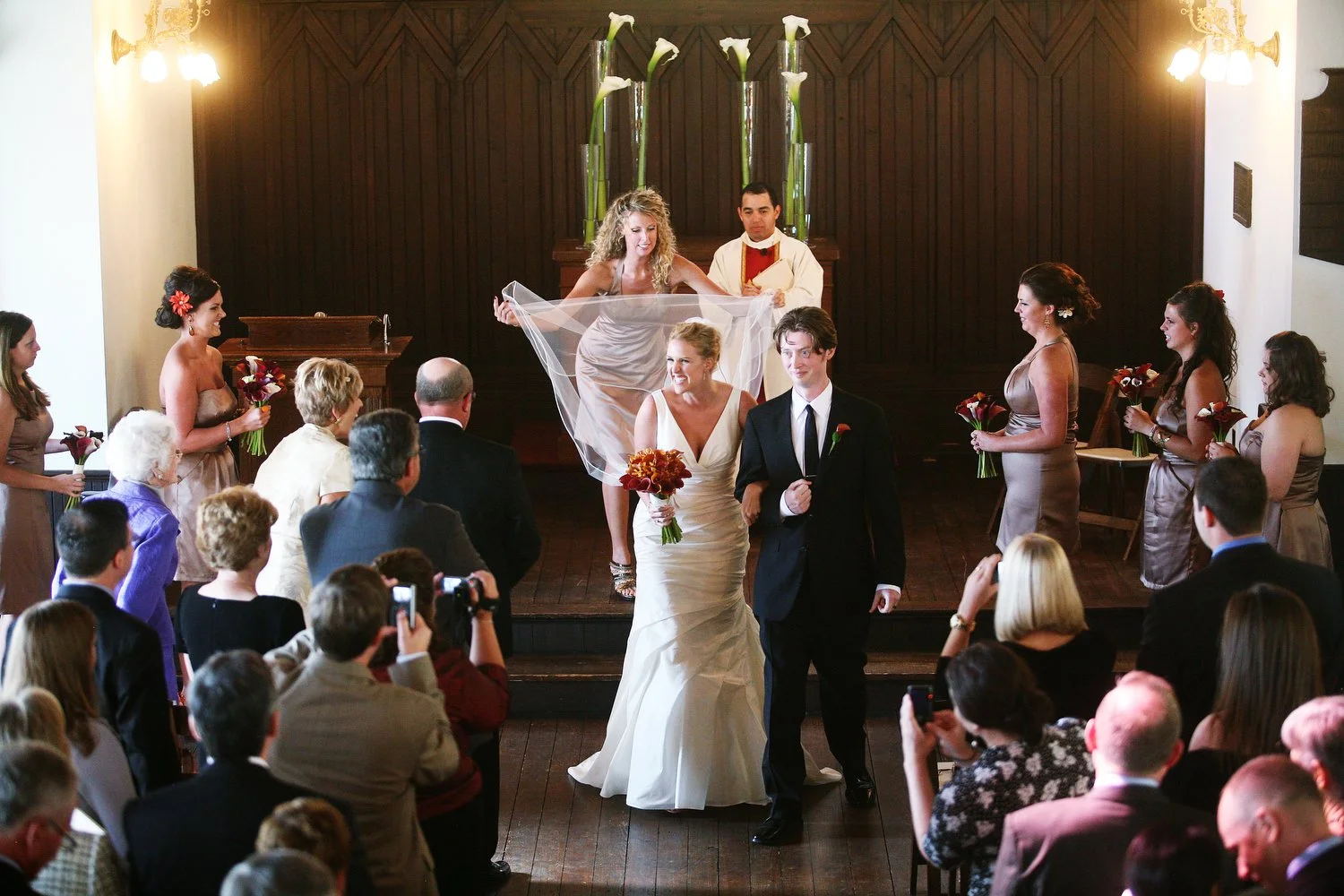 Bride and groom walking down the aisle after their wedding ceremony, with bridesmaids and guests watching. The bride has her chapel length veil straightened behind her by a bridesmaid. 