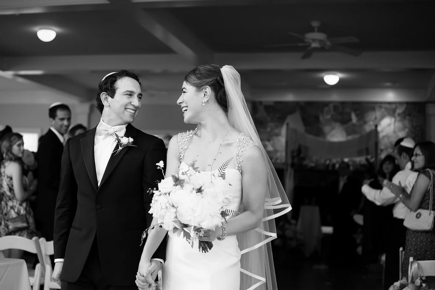 Black and white photo of a bride and groom smiling at each other as they exit their wedding ceremony. He is wearing a kippah. She has a long white veil and is holding a bouquet of flowers. Wedding guests can be seen behind them.  
