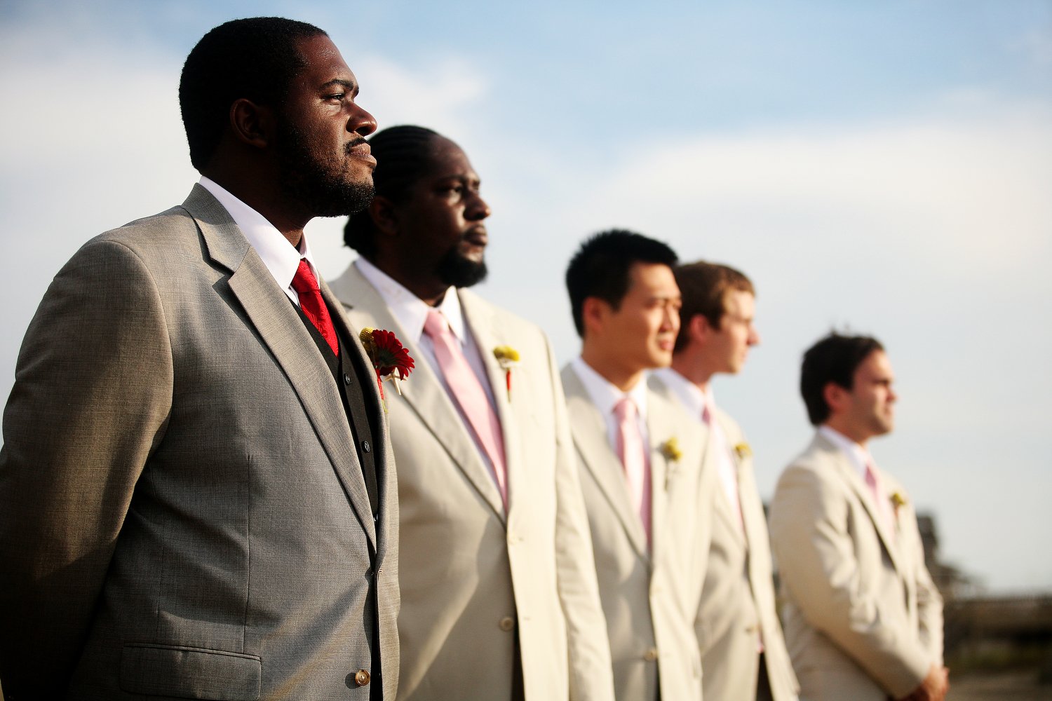 A group of five men in suits standing outdoors, looking to the left, with a cloudy sky. The groom has a grey suit with a red tie. The groomsmen have white suits.