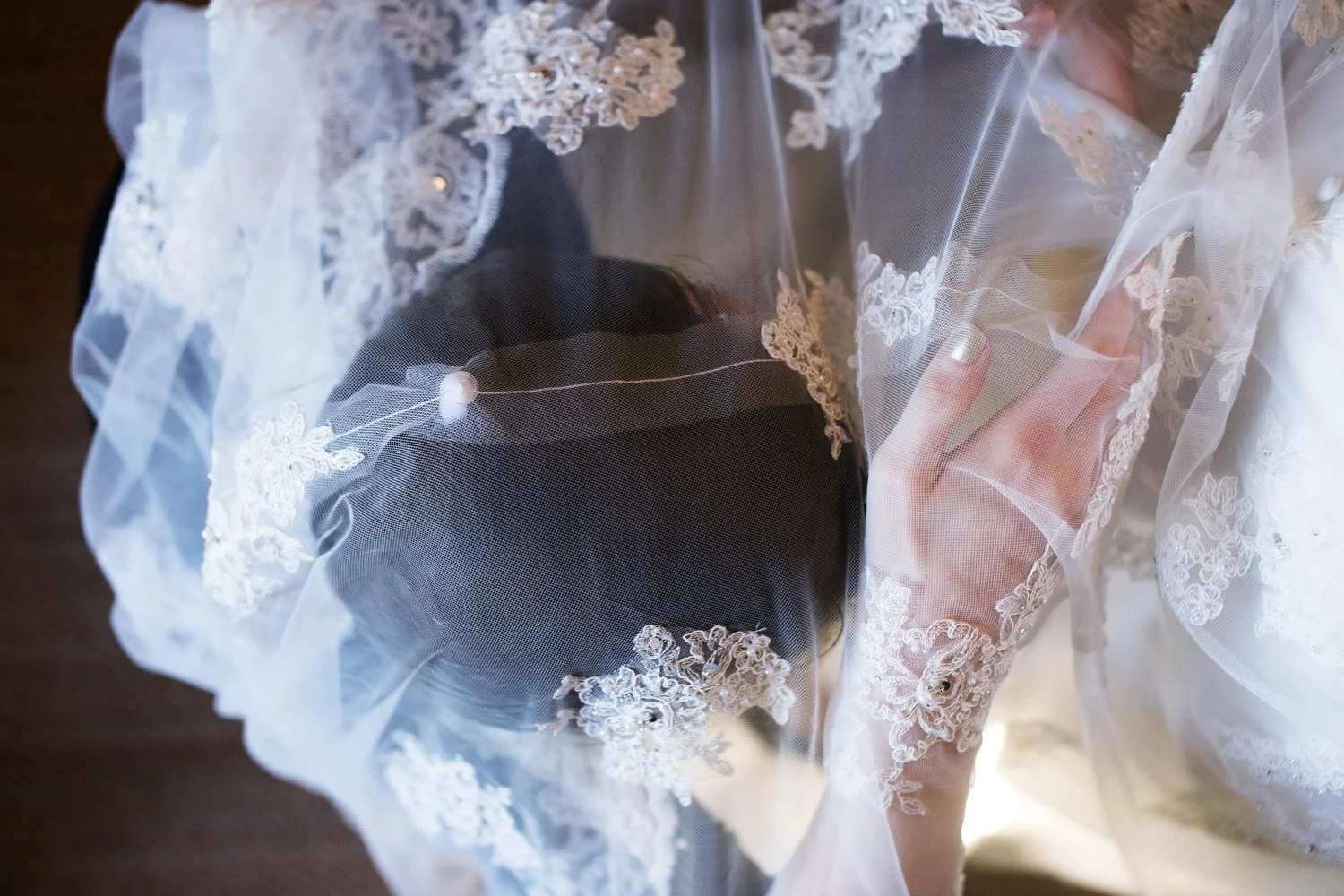 A bride adjusts a sheer lace veil over her head. Only the top of her head is visible through the delicate fabric.