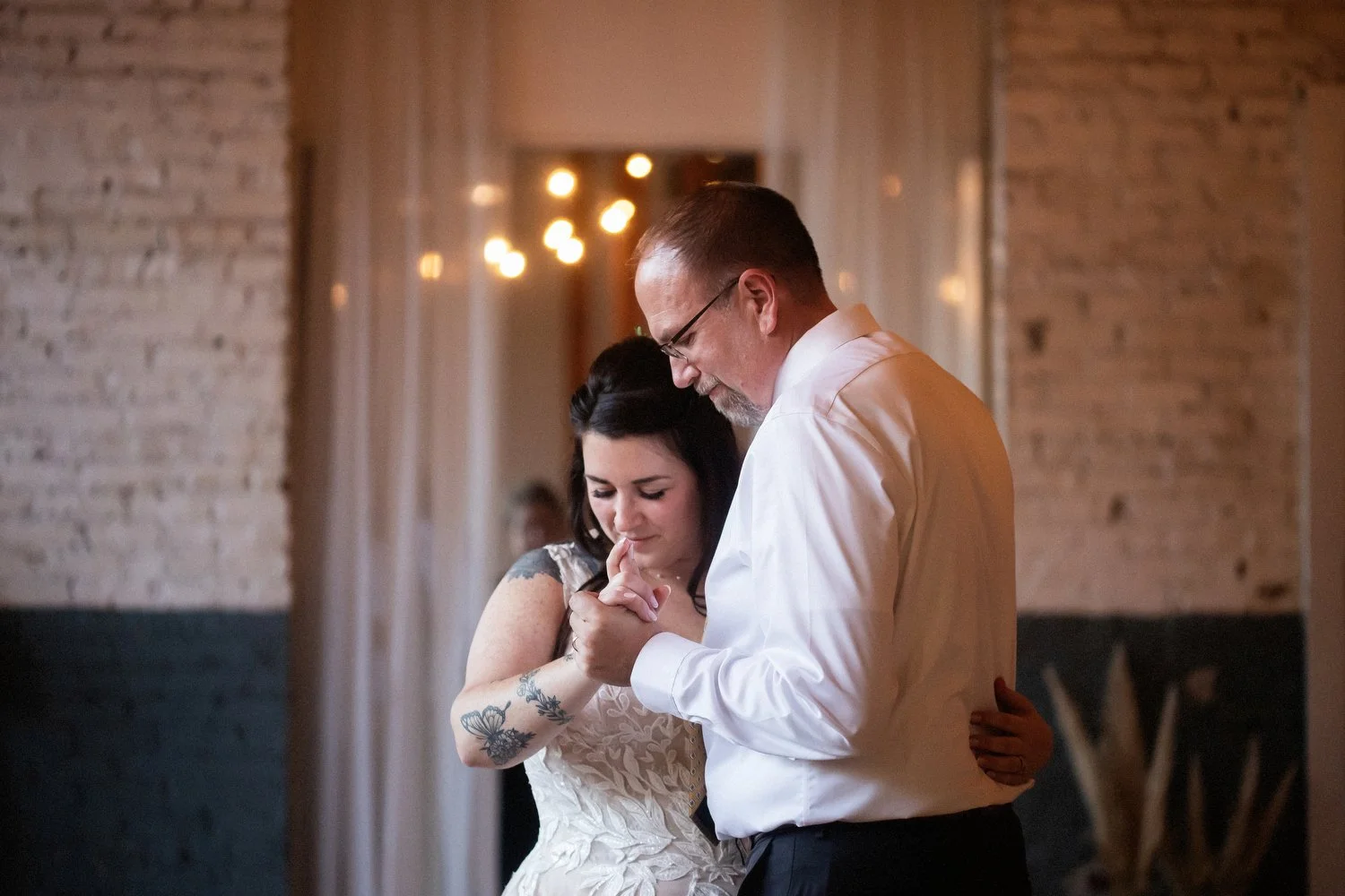 A bride shares a dance with her father at a wedding reception. She is in a lace dress and visible tattoos on her arm, and the man is in a white shirt with glasses, in a warmly lit indoor setting.