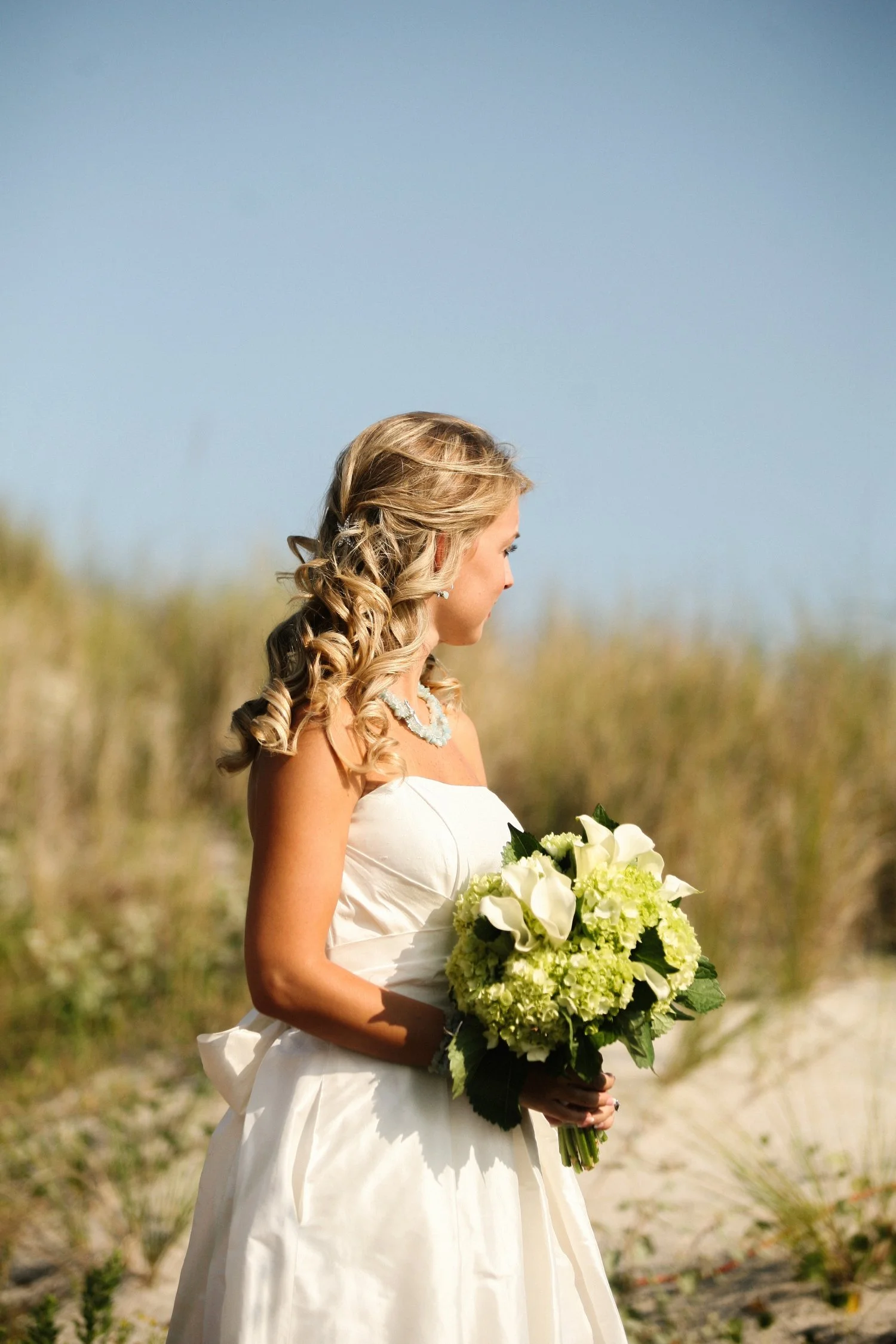A bride in a white wedding dress holding a bouquet of white and green flowers on a sandy beach with grassy dunes and blue sky in the background.