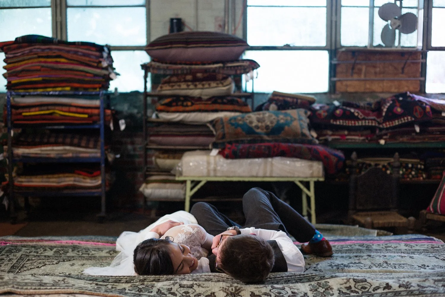 A bride and groom lie down on a huge decorative rug facing each other. She has a white dress with sheer long sleeves. He has on a white dress shirt and vest with grey pants. There are pillows, rugs and other textiles lined up on shelves in front of g