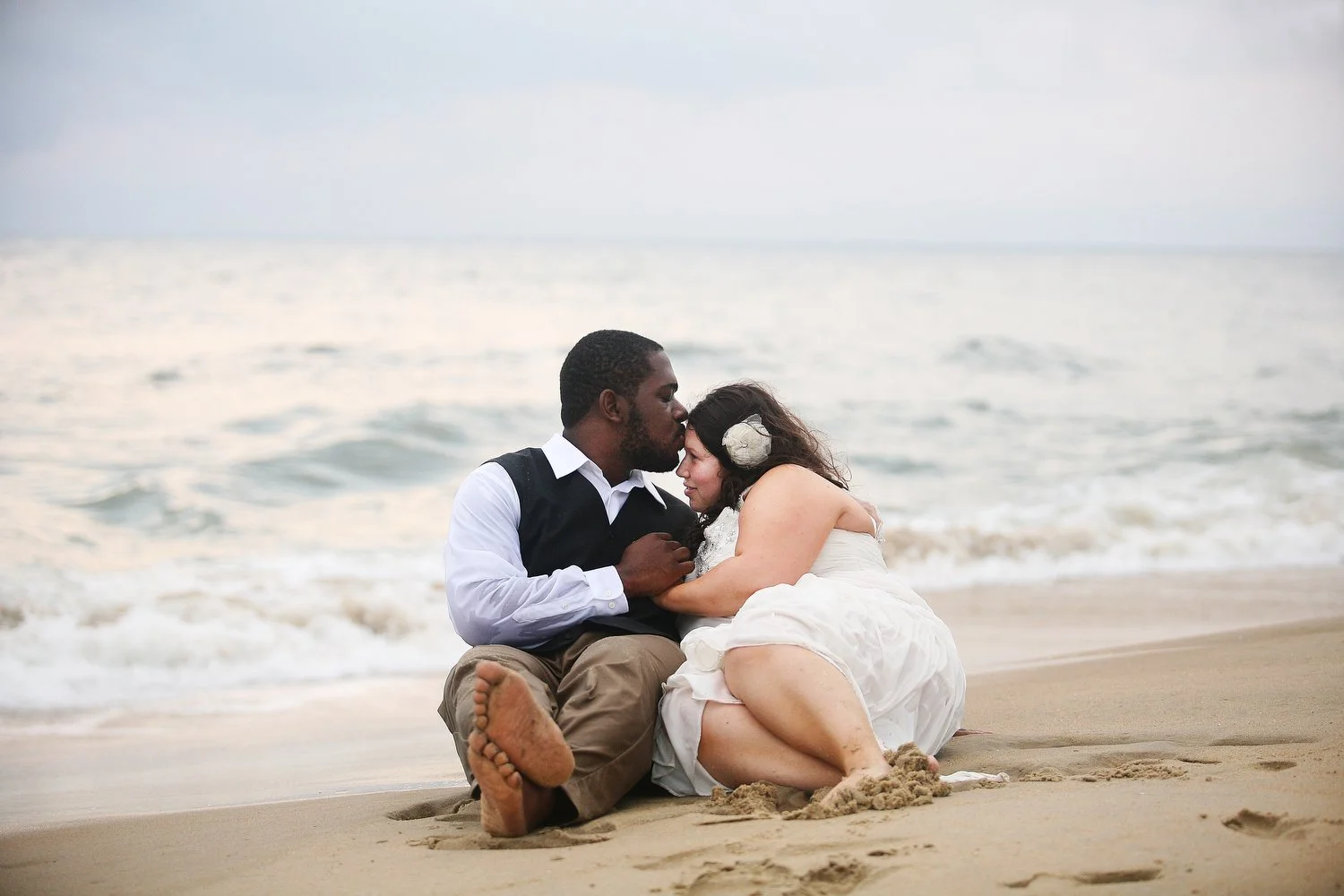 A couple sitting on the beach, with the man kissing the woman's forehead, both embracing each other near the ocean waves in their wedding attire. She has a white dress and a flower in her hair. He is wearing a dressed-down version of a wedding suit. 