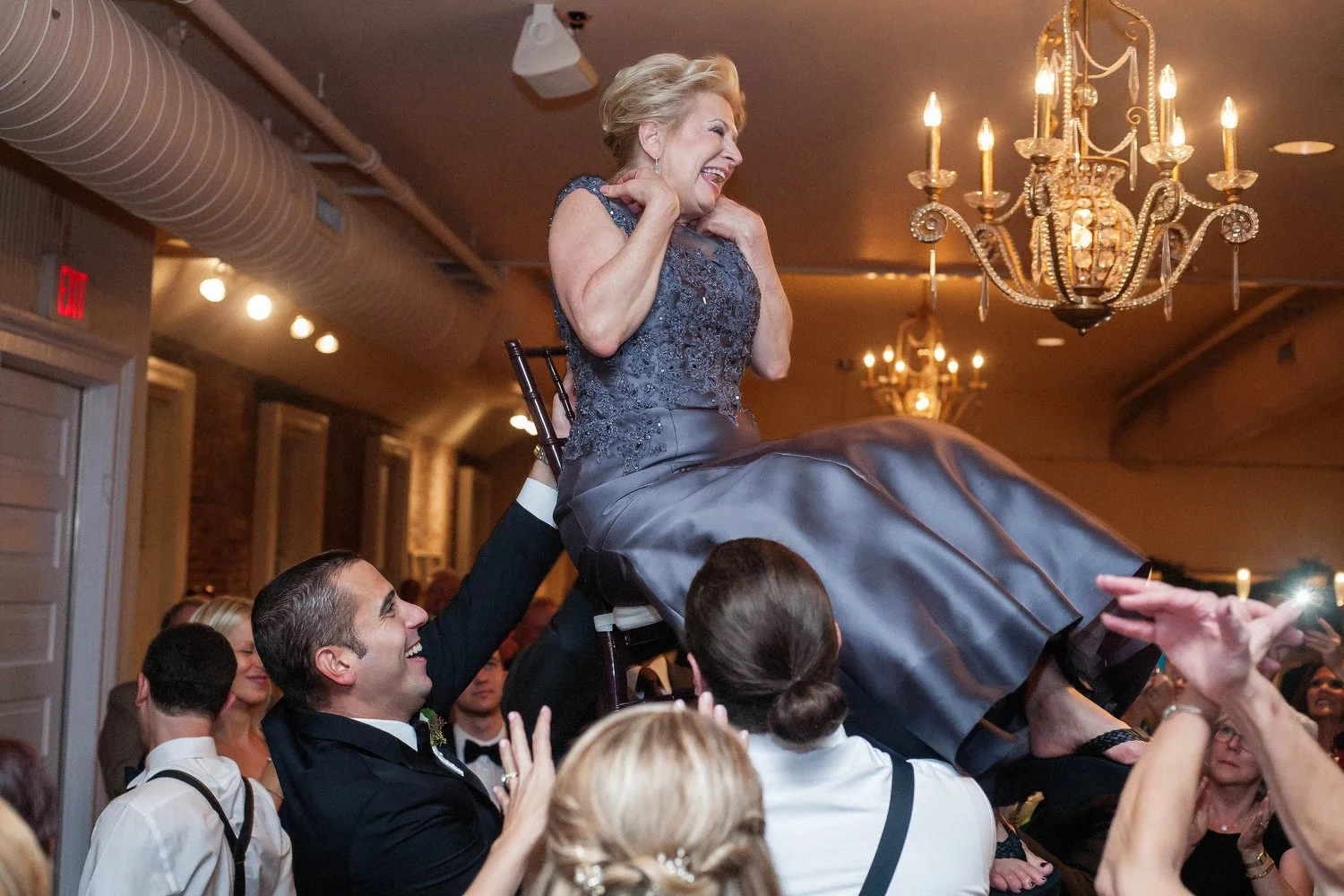 A woman in a formal dress being lifted on a chair by a man at a celebration or wedding reception, with others smiling and clapping around them in a decorated venue with chandeliers.