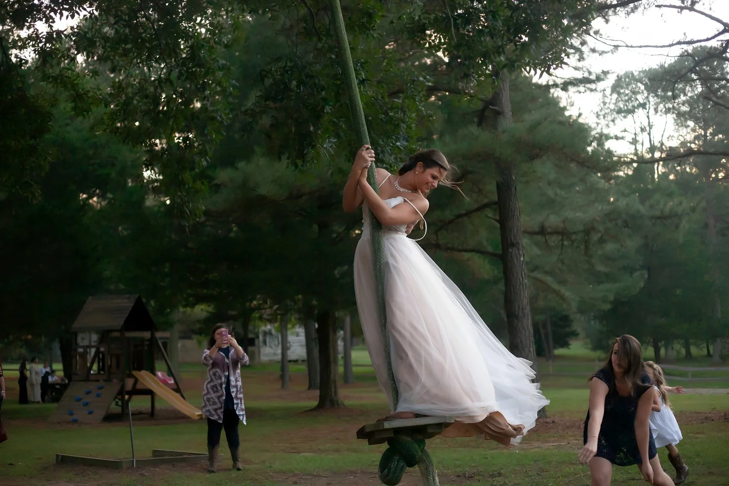 A woman in a white dress is swinging on a large rope swing in a wooded setting, with her feet off the ground and smiling. There are children and adults in the background.