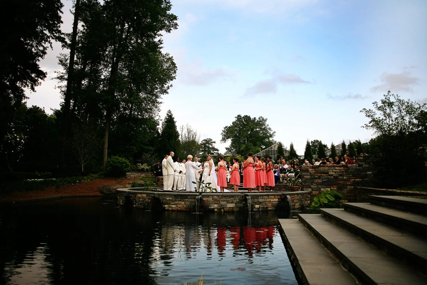 A wedding ceremony taking place outdoors around sunset, with a bride and groom standing in front of a group of bridesmaids dressed in coral pink dresses and groomsmen dressed in white suits, on a raised stone platform beside a pond with reflections, 