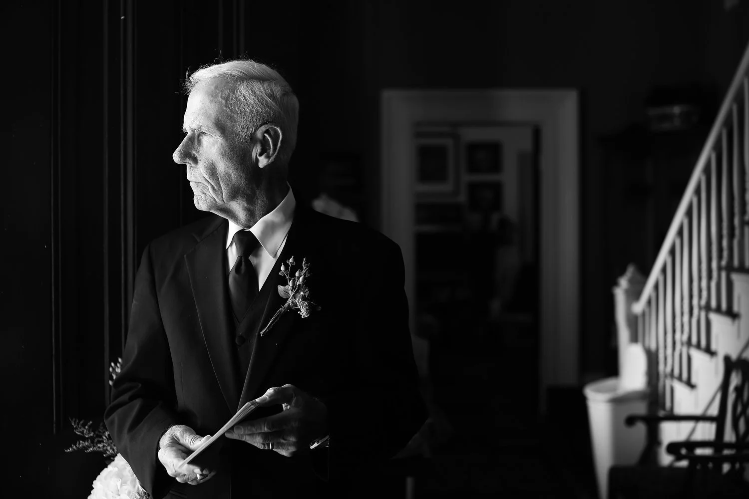 Black and white photo of an elderly man in a suit with a boutonniere, standing indoors and looking to the side while holding a small book or pamphlet. Natural light is on his face.