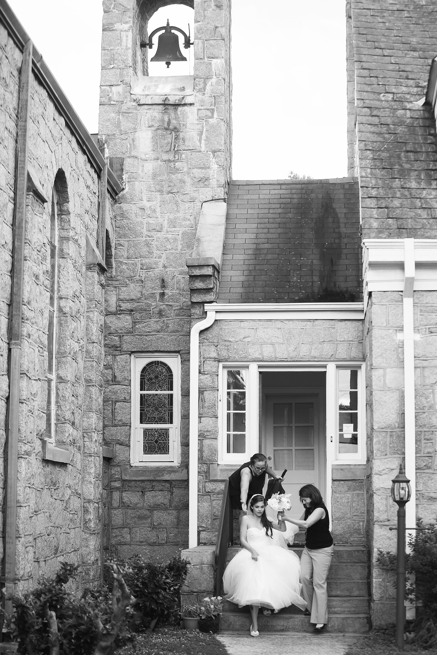 Black and white photo of a bride walking down stairs being assisted by two women, one adjusting her dress and the other holding a bouquet, outside a stone church with a bell tower.