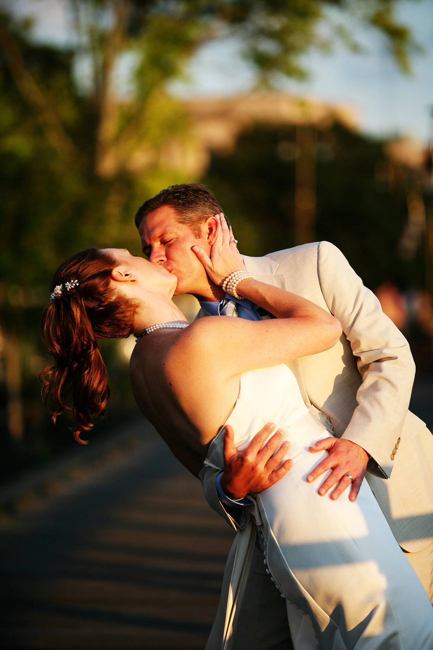 A newlywed couple sharing a kiss outdoors during sunset, with palm trees and a dock path in the background.