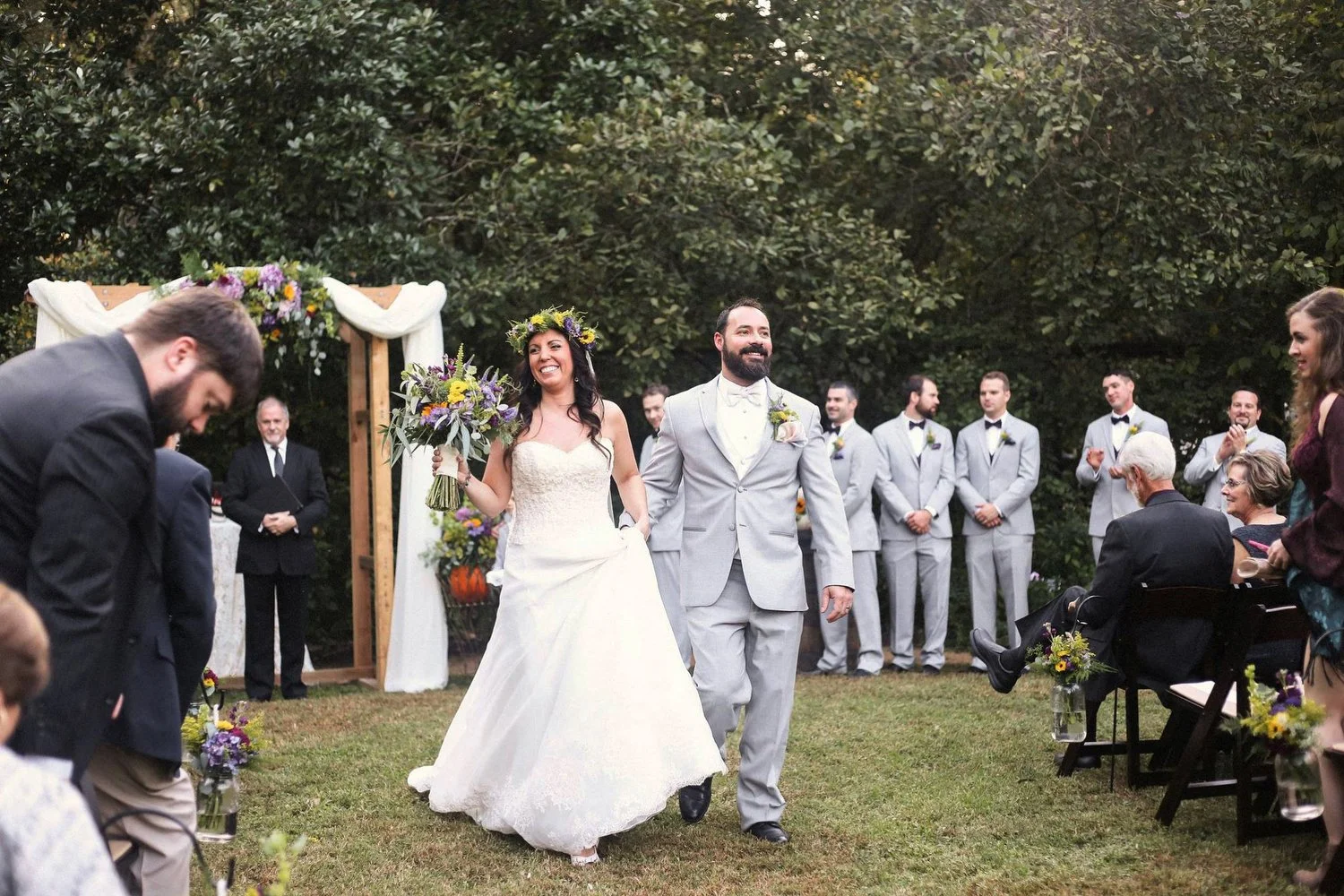A bride and groom walk down the aisle at an outdoor wedding. The bride is holding a bouquet and wearing a white wedding dress with a floral crown. The groom is dressed in a light gray suit with a bow tie and boutonniere. 