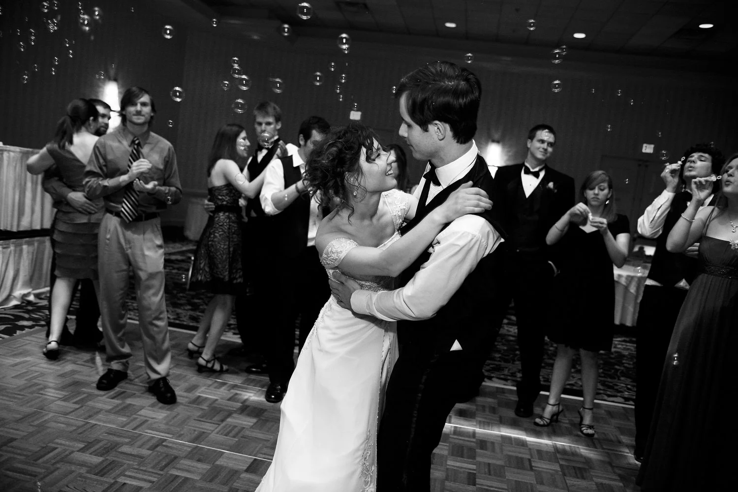 Black and white image of a bride and groom dancing while guests blow bubbles around them. She has her arms up around his shoulders and is leaning in and looking up at him smiling. He has his arms around her waist at the small of her back. 