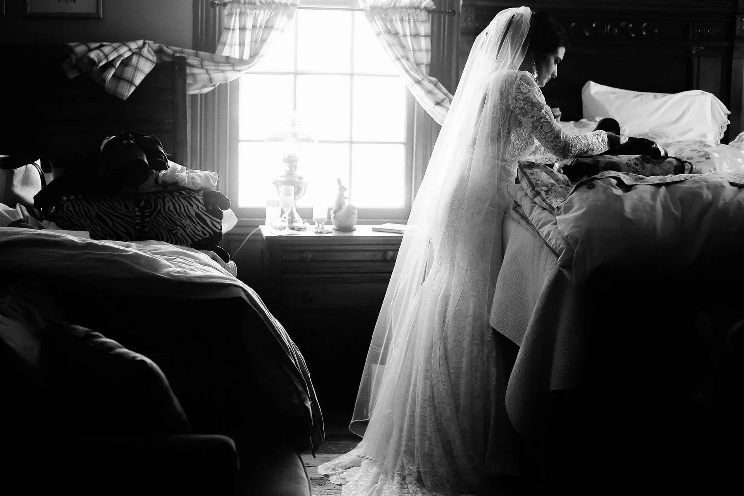 A woman in a long-sleeved lacy wedding dress with a floor-length veil stands next to a bed, looking at a something. There is another bed behind her on the other side of the room. The rustic room has a window with sheer curtains giving the room light.