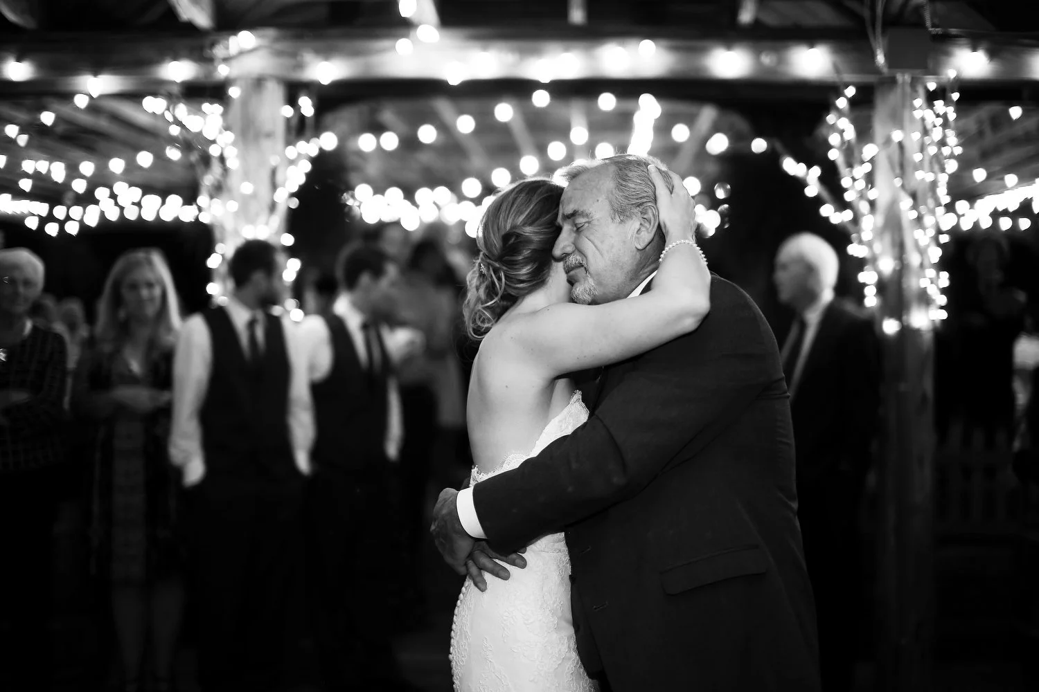 Black and white photo. Tons of twinkle lights illuminate the background and ceiling. A bride leans into her father and rests her right cheek on his right cheek and reaches up with her right arm to clasp the back of his head. He leans in emotionally a