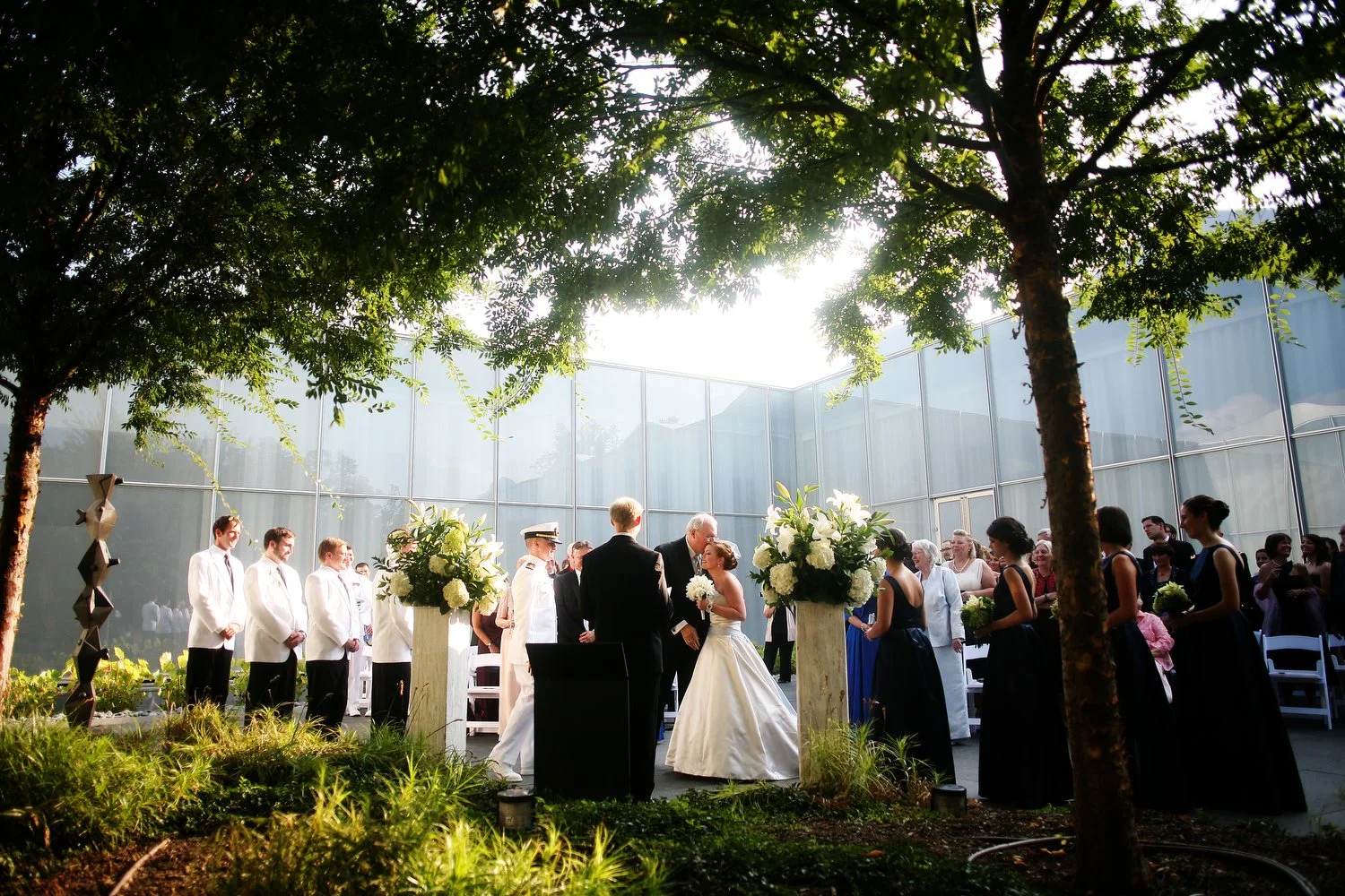 A wedding ceremony taking place outdoors under trees with sunlight shining through, featuring a bride and groom, surrounded by their wedding party and guests. The bride's father is kissing her as he prepares to walk to his seat. The building behind t
